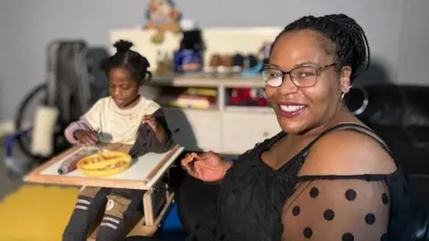 A black woman in a black lacy top sits next to a little boy sitting in a high chair eating a meal.  