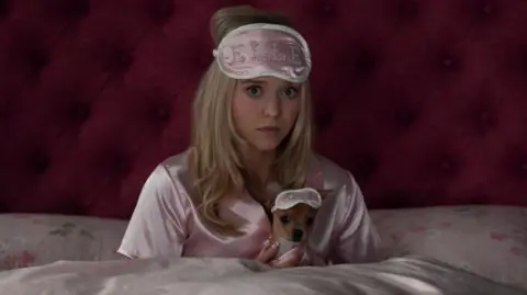 Kimberley French/Prime Video A woman with long blonde hair sits up in bed, with pink bedsheets, pink satin PJs and a pink eye mask on her forehead that has "ELLE" written in diamontes. She's holding a tiny dog which also has a little eye mask on.