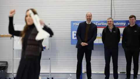 PA Media Prince William and two staff members stand in a line from afar on the curling rink watching Catherine who is not in focus and cheering with her hands in the air.