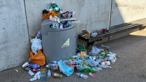 The New Brighteners A grey public waste bin overflowing with rubbish