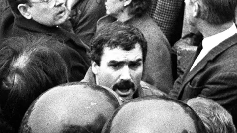 PACEMAKER PRESS A black and white photo of people. One mans face is visible, he has short black hair and a black moustache. Half of one other mans face is visible, he is wearing glasses and has a moustache. 