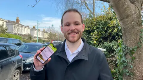 Councillor Izaac Tailford smiling holding his phone on a residential road in Cheltenham with the Litterlotto app open