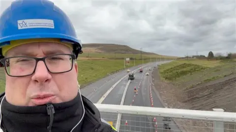 BBC Wales Journalist Pete Shuttleworth in front of the heads of the valleys road works. He is wearing a helmet. 