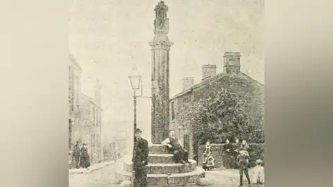 Kirklees Council A black and white photograph of two men standing by a stone cross in Victorian fashion. There are houses to the left and right of the cross