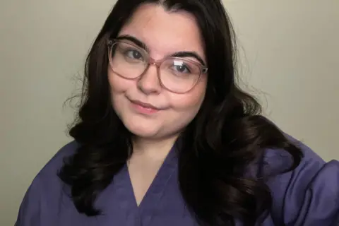 Ayla Jones Head and upper body shot of Ayla Jones, who is white and has long dark slightly wavy hair, and is wearing large, light-brown framed glasses. She has a purple medical scrubs top on and is smiling with her head tilted to her left, looking to the left of the camera.