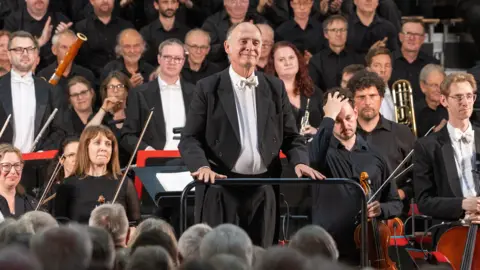 A conductor stood in front of an orchestra addressing the crowd during a performance of the Glagolitic mass at Gloucester Cathedral during the Three Choirs Festival. He is wearing a black suit with a white shirt and is smiling. He is surrounded by an orchestra.