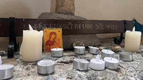 BBC Two white pillar candles and 10 tealight candles sit on a marble surface at the base of a religious shrine.
