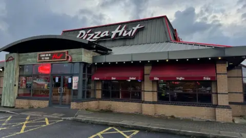 BBC The exterior of a Pizza Hut restaurant with a grey roof displaying a large 'Pizza Hut sign' and maroon window awnings on the windows.