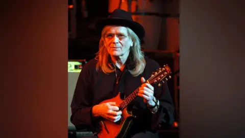 Brian Rasic/Getty Images Henry McCullough Of Slim Chance, playing in 2004. He is dressed in black and holding a mandolin.He is wearing a black hat