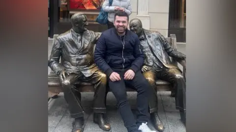 Arlind Xhokola is wearing a navy bomber jacket and smiling. He has short black hair and a beard. He is sitting on a bench with bronze statues on both sides of him.