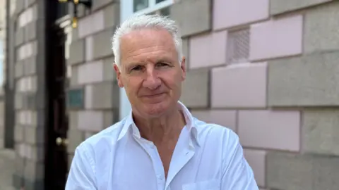 Tom Binet stands outside a stone-bricked building. He has short grey hair and is wearing a white shirt.