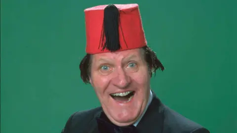 Tommy Cooper is in the middle of the picture, smiling. The picture is taken from above the shoulders, and he is wearing a black suit jacket, and bow tie. He is wearing a fez, which is a red hat with a black tassel on. 