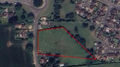 Google Aerial view of fields and houses with a red line demarking the area of the proposed development.