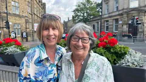 A woman wearing a flowery patterned blouse, stands on the left, she has a bob haircut and sunglasses on her head. To her right stands an older lady with a grey bob haircut, she is wearing a white blouse. Behind them are flower baskets and Pudsey Town Hall is behind them.