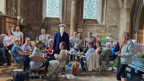 Rheged A large group of women sitting in a church hall working on their panel. There is a life-size model of a sailor in the middle.