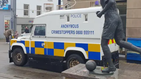 A police van parked in a town centre during the day behind a statue of a footballer. Some people can be seen in the background.