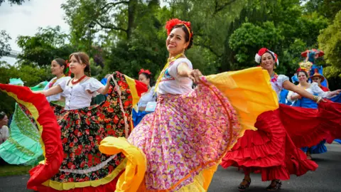 Pacemaker Press Women are dressed in colourful dresses ad dancing in a festival. Some have flowers in their hairs. Trees are behind them.