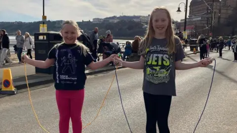 Bek Homer/BBC Two girls stand smiling on the Scarborough Foreshore holding skipping ropes. 