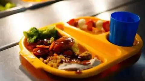 A yello compartmentalised tray containing a school meal of meat and vegetables with a blue cup