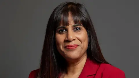 The MP Sureena Brackenridge is looking directly at the camera. The background is grey. She has dark long hair and is wearing a red jacket. 