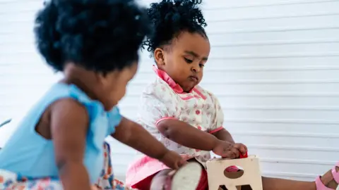 Two  young girls, one with a blue top, one with pink and white floral clothes, playing with a wooden toy.