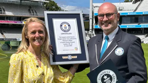 A woman in a yellow summer dress smiles for the camera as she holds up a certificate of achievement from Guinness World Records with an official from the organisation.