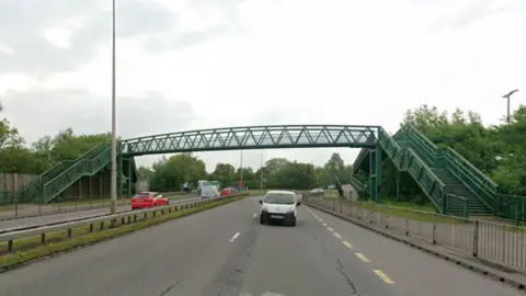 Spytty Road, a multi-lane road runs beneath a green metal pedestrian footbridge with staircases on both sides. Several cars and a white van travel along the road, and trees line both sides.
