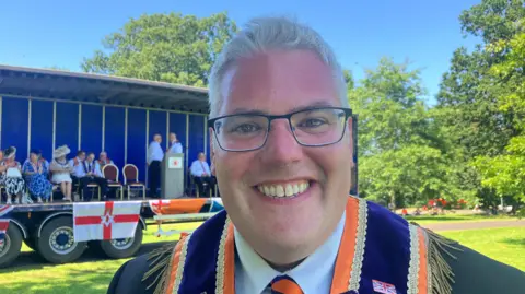 A man with short white hair and black rimmed glasses is smiling and looking into the camera. He is wearing a dark coloured suit jacket and an orange and purple sash.