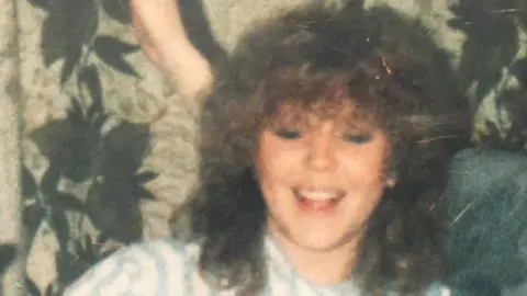 Family Photo Lynette White when she was alive, she has long curly hair and is smiling at the camera. Her shirt is blue and white stripey.