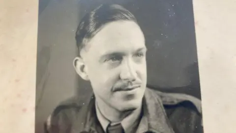 Supplied Leslie Lemon, in an old black and white photo, taken during World War Two, wearing an army uniform, wearing a jacket, shirt and tie. He has slicked back hair and has a moustache.