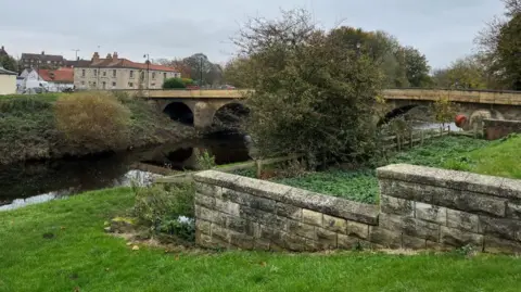 A bridge over a river with grass banks on either side and a stepped brick wall going up the right hand side of the bank