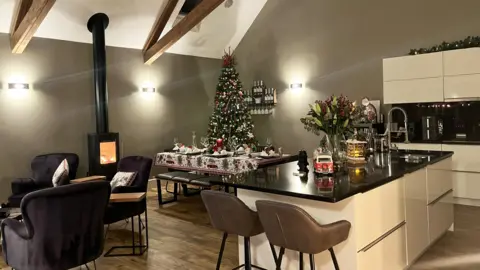 IWC Media Modern kitchen and dining area decorated for Christmas with a large lit tree, festive table setting, and cozy seating near a wood stove.