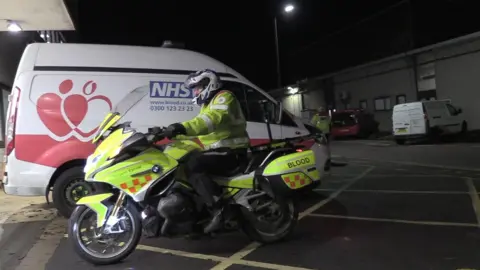 A motorcyclist in a high vis jacket on a high vis motorbike pulling in to park the bike next to an NHS blood van. His bike has "blood" clearly visible on the rear of the vehicle.