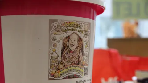 A pink and white charity collection bucket, with a sticker of Margo on the front and the name of her charity 'Margo's Marvellous Mind.' 