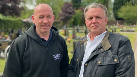 Two men, one bald, the other with longer blonde and grey hair, which is swept back. The bald man wears a black top with the words Nettlebank Memorials Limited on the chest. There are gravestones behind them