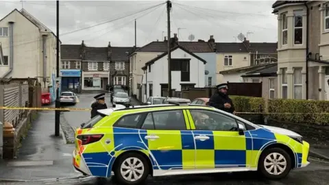 Bishop Road blocked by a police cordon. A police car is in the foreground of the picture. There are two police officers in uniform next to the car. Yellow cordon tape is also across the street at both ends. It is a grey day and shops at the end of the street are closed.