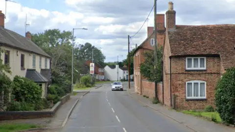 Google A village street with old houses and trees down both sides and a car in the distance