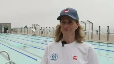 A woman wearing a white t-shirt and blue hat stood in front of a swimming pool. The pool is next to a rocky beach, with the sea in the background.