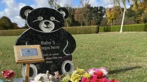 Sackers A large bear-shaped headstone with the Baby S written on it. It is positioned in a field and in front of some flowers and a wooden and mental plaque
