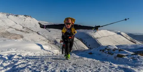 David Murch/Montane Spine Race A man puts his hands out in celebration after reaching the top of a snowy hill. He has dark glasses, a bald head and a yellow backpack.
