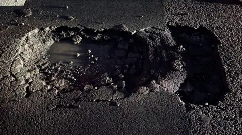 A close-up view of a damaged section of asphalt road surface. At the centre of the image, there is an irregularly shaped pothole with rough, crumbling edges where the top layer of the road has broken away. The inside of the pothole appears dark and damp. Loose stones and fragments of asphalt are visible around the edges of the hole.