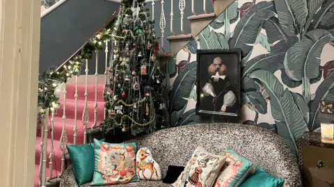 IWC Media Stylish hallway with a Christmas tree by a staircase, a patterned sofa with festive pillows, and bold leaf-print wallpaper. On the wall behind the sofa is a painting of a poodle in a medieval dress.