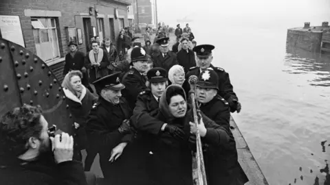 Mirrorpix/Getty Images A group of policemen surround and attempt to detain a woman with dark hair wearing a headscarf - she holds onto the metal barrier of a dock. More people can be seen on the dock, with water off the side. There is a man taking photographs of the kerfuffle in the foreground.