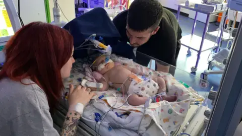 Tia Tia with dark red hair sits beside a hospital cot, her tattooed arm reaching towards the baby in the middle. Arabella is wearing a nappy, on a ventilator and has wires and tubes attached to her. Blaze is stood being over looking down at her on the other side of the cot. Medical trolleys and paraphernalia visible in the background.