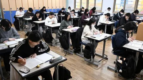South Korean students seated in neat rows in a classroom in Seoul as they wait to take the annual College Scholastic Ability Test, known locally as Suneung, on 14 November, 2024. Several are looking at their mobile phones while others are scribbling on books. 