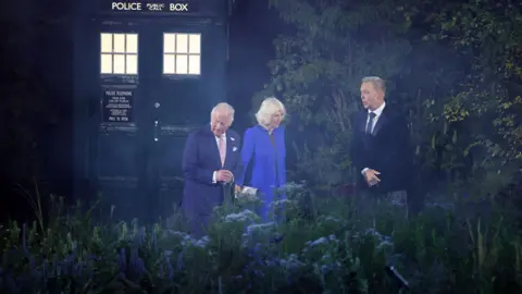 Getty Images King Charles and Queen Camilla walk through the garden, after emerging from a Tardis. Sir Chris Hoy looks on.