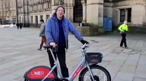 David Robison A man in a blue jacket a beanie hat holds a grey and red e-bike outside Bradford City Hall