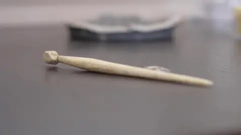 A close-up image of a Roman hair pin made of bone. It is a long, thin pin with a round piece on the end.