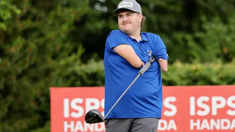 Adam Dalton wearing a blue shirt tucks a golf club under his armpit