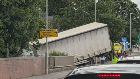 A lorry hangs off the edge of a road at a bridge as traffic builds. Someone in a hi-visibility vest is holding their hand up, directing traffic. There are trees lining the road as well as signs for businesses.